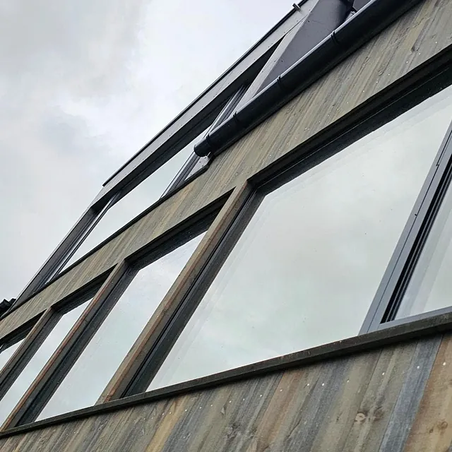 Høy fasade med store vinduer og værbitt trekledning, sett nedenfra.